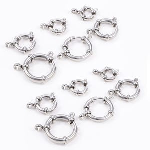 May include: A collection of silver-colored round spring ring clasps in various sizes. Each clasp features a circular design with a hinged opening and a small lever for secure closure. The clasps are arranged on a white surface.