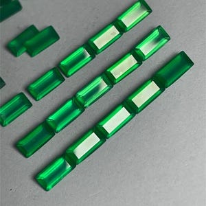 May include: Bright green rectangular gemstones with a polished finish. The gemstones are arranged in rows and scattered across a gray surface. The gems have a translucent quality, with a lighter center.