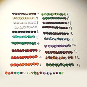 May include: A selection of small, heart-shaped gemstones in a variety of colours, arranged in rows and numbered. Colours include red, orange, green, black, and clear. The gemstones are displayed on a white surface.