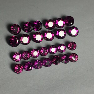 May include: A collection of 20 round, faceted, purple gemstones. The gemstones are arranged in rows on a gray surface.