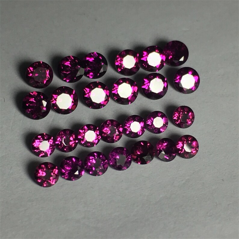 May include: A collection of 20 round, faceted, purple gemstones. The gemstones are arranged in rows on a gray surface.