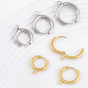Puede incluir: Una variedad de pendientes de aro en tonos plateados y dorados. Los pendientes varían en tamaño y diseño, algunos con un cierre de bisagra. Se exhiben sobre una superficie blanca, mostrando su acabado pulido y su elegancia sencilla.