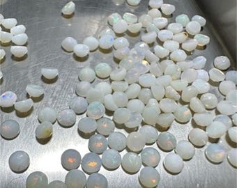 Può includere: Un grande gruppo di cabochon di opale bianco, un tipo di pietra preziosa, sono sparsi su una superficie argentata. Gli opali sono rotondi e hanno una finitura liscia e lucida.