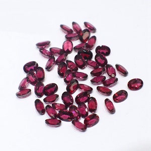 Può includere: Una collezione di pietre preziose di forma ovale di colore rosso scuro. Le pietre preziose sono sparse su una superficie bianca.