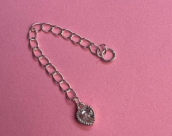 May include: A silver-colored chain extender with a heart-shaped charm. The chain is composed of oval links and has a small ring at one end. The heart charm is encrusted with small, clear stones. The background is a solid pink color.