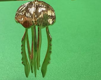 May include: A gold-colored jellyfish pendant with a textured cap and flowing tentacles. The pendant is suspended from a thin, curved gold bar. The background is a solid green.
