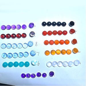 Peut inclure: Une collection de pierres précieuses rondes et colorées disposées en rangées. Les pierres précieuses varient en couleur, notamment des nuances de violet, de rouge, d'orange, de bleu et de blanc. Chaque pierre mesure environ 1,3 cm de diamètre. L'image est prise d'en haut.