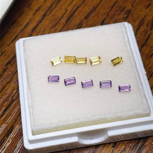 Puede incluir: Una bandeja blanca contiene varias piedras preciosas rectangulares. La fila superior presenta gemas amarillas, mientras que la fila inferior muestra gemas moradas. Las gemas se exhiben sobre una superficie blanca, sobre un fondo de madera.