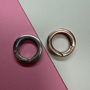 Puede incluir: Dos cierres redondos de metal, uno plateado y otro oro rosa, se muestran sobre un fondo rosa y blanco. El cierre plateado tiene el sello "925". Estos cierres se utilizan probablemente para la fabricación de joyas o manualidades.