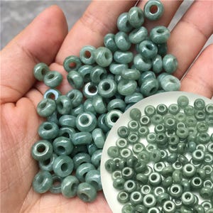 Peut inclure: Un ensemble de perles en forme de donut de couleur vert jade. Les perles sont d'un vert clair et ont une surface lisse et polie. Certaines perles sont dans une main, d'autres dans une petite coupelle blanche.