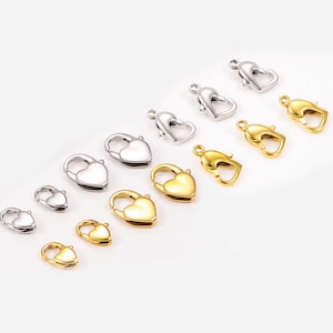 Könnte beinhalten: Eine Auswahl an herzförmigen Karabinerverschlüssen in Silber- und Goldtönen. Die Verschlüsse sind diagonal angeordnet und zeigen verschiedene Größen und Ausführungen. Diese Schmuckzubehörteile eignen sich ideal für Halsketten, Armbänder und andere handgefertigte Bastelarbeiten.