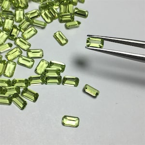 Può includere: Una collezione di pietre preziose di peridoto verde in taglio smeraldo rettangolare. Le pietre sono sciolte e sparse su una superficie bianca. Una pietra è tenuta da una pinzetta.