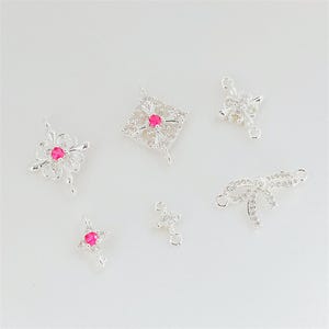 Può includere: Una collezione di sei connettori per gioielli color argento con pietre di zirconia cubica trasparenti. Il set include motivi floreali, a rombo, a fiocco e a stella, con tre pezzi impreziositi da una pietra centrale rotonda rosa acceso.