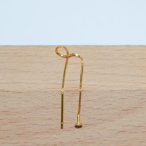 Peut inclure: Une paire de boucles d'oreilles pendantes dorées. L'une des boucles d'oreilles est une barre droite, l'autre un petit clou rond. Les boucles d'oreilles sont présentées sur une surface en bois brun clair.