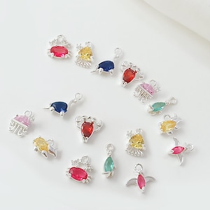Puede incluir: Una colección de pequeños dijes plateados con detalles de piedras preciosas de colores. Los dijes presentan varias formas, incluyendo cangrejos, pájaros y diseños abstractos, con piedras rojas, amarillas, azules, rosas y verdes. Estos dijes son probablemente para la fabricación de joyas.