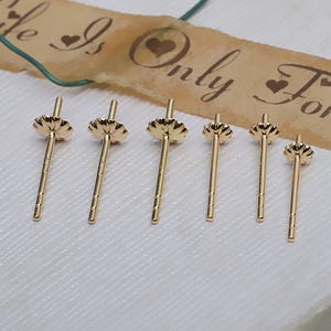 Peut inclure: Six tiges de boucles d'oreilles dorées avec un dessus en forme de fleur et une tige filetée. Les tiges sont disposées sur une surface blanche texturée. Une bande de papier décorative avec les mots "Life Is Only For" est en arrière-plan.
