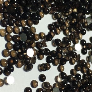 以下が含まれることがあります： 小さな丸いダークブラウンのファセットカットガラスビーズの山をクローズアップした画像。ビーズは白い表面に散らばっています。
