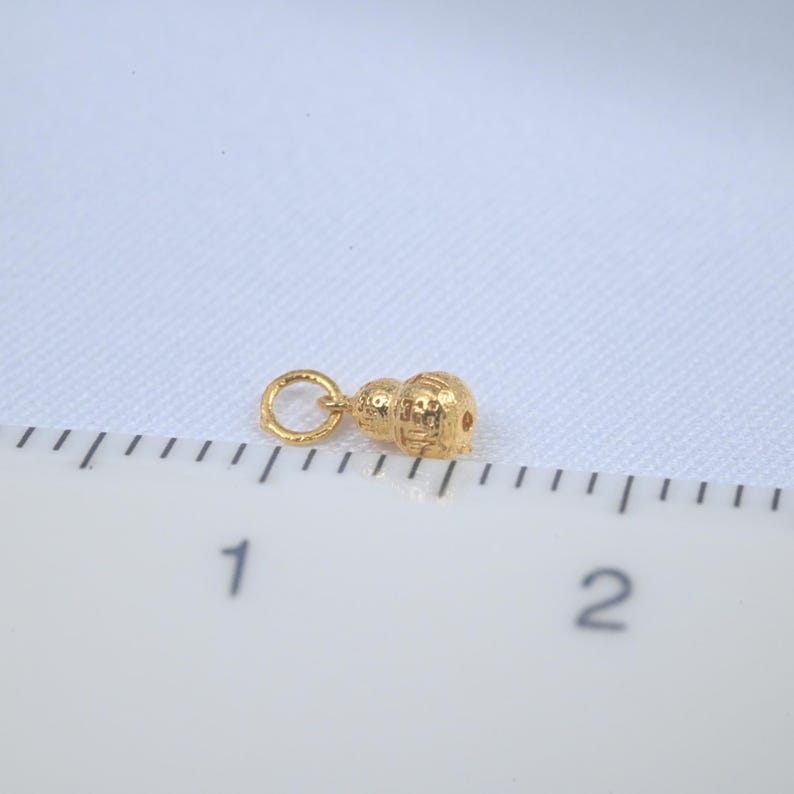 Op de afbeelding: Een kleine, goudkleurige bedel in de vorm van een kalebas. De bedel heeft een getextureerd oppervlak en een kleine lus om aan een ketting of armband te bevestigen. De bedel is ongeveer 1,3 cm lang.