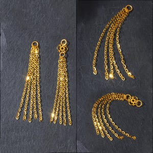 May include: Gold-coloured chain earrings with multiple strands, displayed against a dark background. The earrings have a delicate, dangling design, with a small loop at the top for attachment. The chains are arranged in a cascading pattern.
