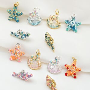 Op de afbeelding: Een verzameling kleurrijke emaille bedels in verschillende vormen, waaronder zeesterren, schelpen en vissen. De bedels hebben goudkleurige metalen accenten en zijn beschilderd in tinten blauw, oranje, roze en groen. Deze decoratieve bedels zijn ideaal voor het maken van sieraden.