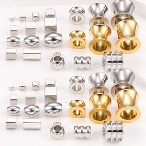May include: A selection of shiny metal jewellery components, including various shapes of beads, plugs, and spacers. The components are in silver and gold tones, with cylindrical, square, and rounded designs. The image showcases a variety of sizes and styles.