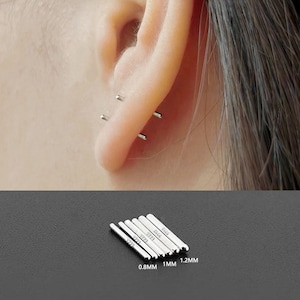 May include: Silver bar earrings in various sizes are shown, with some worn in an ear. The earrings are labeled with their sizes: 0.8mm, 1mm, and 1.2mm. The earrings are small and minimalist, suitable for multiple piercings.
