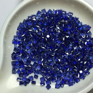 Puede incluir: Una vista cercana de una colección de pequeñas piedras preciosas azules de forma cuadrada. Las piedras preciosas están esparcidas sobre una superficie blanca.