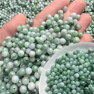 May include: A close-up of a collection of light green jade beads. The beads are round and vary in shade, with some appearing more opaque and others translucent. Some beads are held in a hand, others in a white bowl, and the rest are scattered.