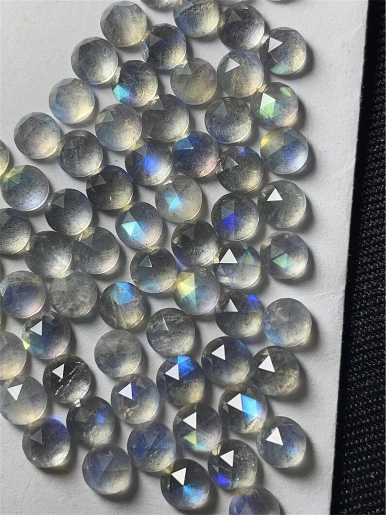 May include: A collection of iridescent, faceted, round labradorite gemstones. The stones are a light gray color with blue and green iridescence.