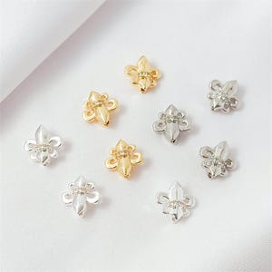Può includere: Una collezione di piccoli ciondoli decorativi a forma di giglio, in tonalità oro e argento. Ogni ciondolo presenta un elemento centrale a punta con tre petali curvi e una piccola gemma trasparente. Su sfondo bianco.
