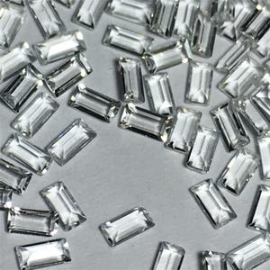 Könnte beinhalten: Eine große Menge an klaren, rechteckigen, facettierten Edelsteinen. Die Edelsteine sind auf einer weißen Oberfläche verstreut.