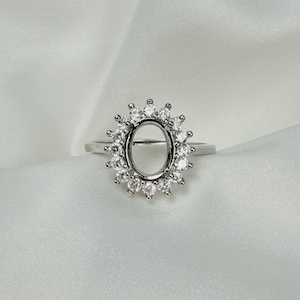 May include: A silver ring setting with an oval center and a halo of small, round, clear gemstones. The ring is set against a white, textured background, highlighting the ring's design and sparkle.