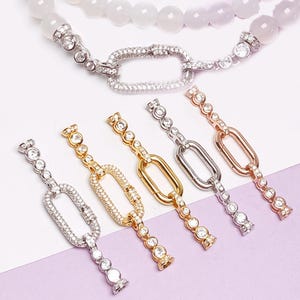May include: An assortment of jewelry components, including silver, gold, and rose gold-toned chain links, each adorned with sparkling clear crystals. A necklace with white and light purple beads is visible in the background. The links are designed for jewelry making.
