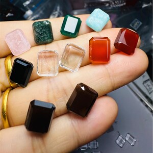 Op de afbeelding: Assortiment van tien gefacetteerde edelsteen-cabochons in verschillende kleuren, waaronder zwart, bruin, helder, groen, rood, roze en blauw. De edelstenen worden op een hand getoond.