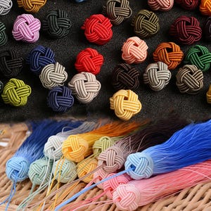 Könnte beinhalten: Eine Sammlung farbenfroher dekorativer Knotenknöpfe in verschiedenen Farbtönen, darunter Rosa, Rot, Grün und Gelb. Die Knöpfe bestehen aus gewebtem Kordel und werden mit passenden Quasten präsentiert.