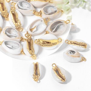 May include: A selection of white cowrie shell charms with gold-coloured metal accents. The shells have a natural white exterior, with gold-coloured metal detailing. Each charm has a small loop for attaching to jewellery or other crafts.