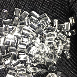 Puede incluir: Un montón de piedras preciosas rectangulares transparentes y facetadas. Las piedras preciosas están esparcidas sobre un fondo de tela negra.