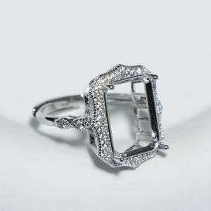 Puede incluir: Un anillo de plata con un engaste de talla esmeralda, incrustado con pequeñas piedras preciosas claras. La banda del anillo presenta detalles delicados y pequeñas piedras preciosas adicionales. El engaste está vacío, listo para una piedra preciosa grande.