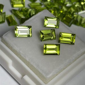 Puede incluir: Una colección de ocho piedras preciosas de peridoto verde de forma rectangular. Las piedras preciosas se muestran en una superficie de espuma blanca dentro de una caja blanca.
