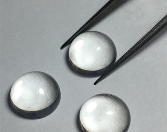 May include: Three clear glass cabochons, each with a slightly different shade of grey, are displayed on a white surface. The cabochons are round and smooth, with a slight dome shape.