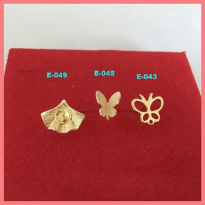 Könnte beinhalten: Drei goldfarbene Metall-Anhänger auf rotem Hintergrund. Die Anhänger sind in Form eines Fächers, eines Schmetterlings und eines Schmetterlings mit einem detaillierteren Design. Die Anhänger sind mit E-049, E-045 und E-043 beschriftet.