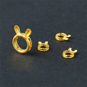 Puede incluir: Cuatro abalorios de metal de color dorado en forma de conejo con orejas. Los abalorios están diseñados de forma intrincada con un patrón delicado.