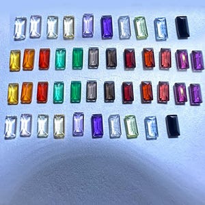 May include: A selection of rectangular gemstones in a range of colours, such as clear, yellow, orange, red, green, grey, brown, and purple. The gemstones are arranged in rows, displaying their different hues and clarity. These gemstones are likely for jewellery making.