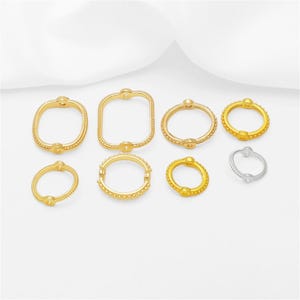 May include: A collection of eight jewellery findings including seven gold-toned metal rings and oval shapes with beaded textures and one smaller silver-toned metal ring. These components feature small circular attachment points along their edges.