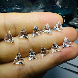Puede incluir: Una colección de 10 piedras preciosas triangulares transparentes, probablemente cuarzo, expuestas en una mano. Las piedras preciosas son facetadas y tienen un ligero brillo.