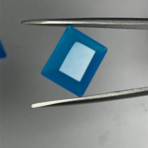 May include: A bright blue gemstone with a rectangular shape, held by tweezers. The gemstone has a clear, white centre.
