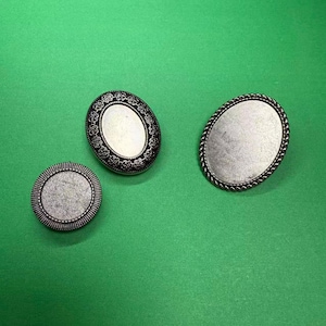 May include: Three silver metal jewellery findings on a green surface. The findings include two oval and one round shape. The oval findings have decorative borders, while the round finding has a dotted border. The centre of each finding is a blank, textured surface.