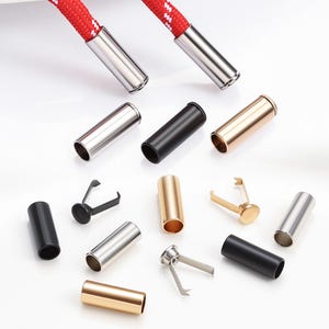 Op de afbeelding: Assortiment van metalen koordeindkappen en koordstoppers in zilver, zwart en goud. Twee rode koorden met witte accenten worden getoond met zilveren eindkappen. De afbeelding toont verschillende stijlen koordeindaccessoires.