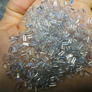 May include: A large pile of clear, iridescent, rectangular gemstones. The gemstones are scattered on a hand.