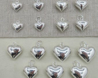 Könnte beinhalten: Eine Sammlung silberner herzförmiger Anhänger mit poliertem Finish. Jeder Anhänger hat oben eine kleine Schlaufe zum Befestigen an einer Kette oder einem Armband. Die Anhänger variieren leicht in der Größe und werden auf einem neutralen Stoffhintergrund präsentiert.